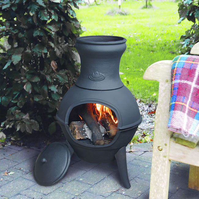 A black chiminea with a glowing fire and wooden logs sits on a patio. A colorful, plaid blanket drapes over a nearby chair, surrounded by lush green plants.